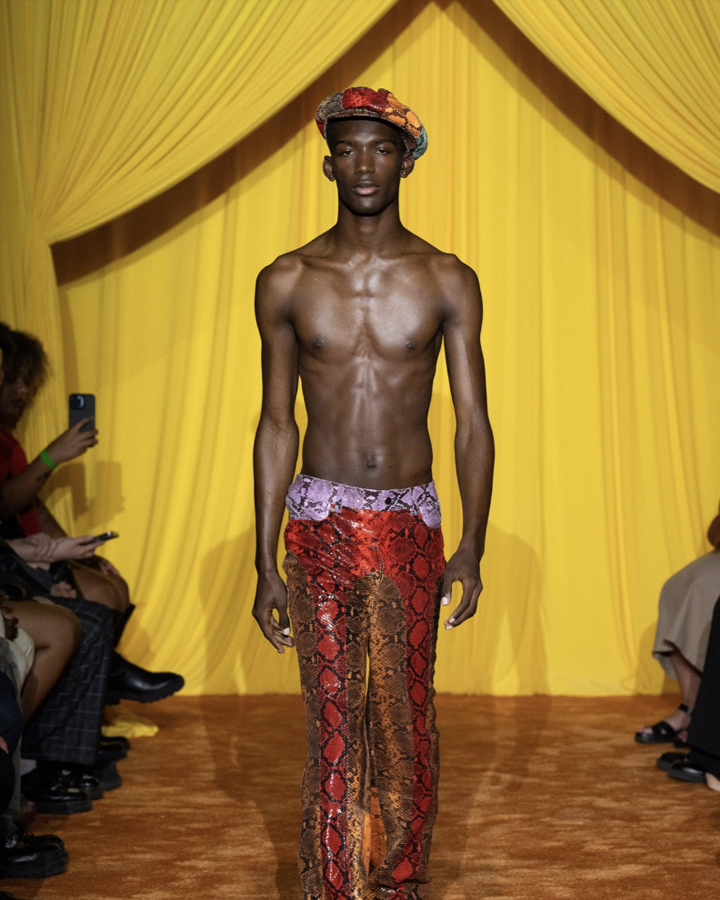 Slender male model with deep skin wearing only snakeskin pants and a snakeskin hat walking on a runway against a yellow backdrop